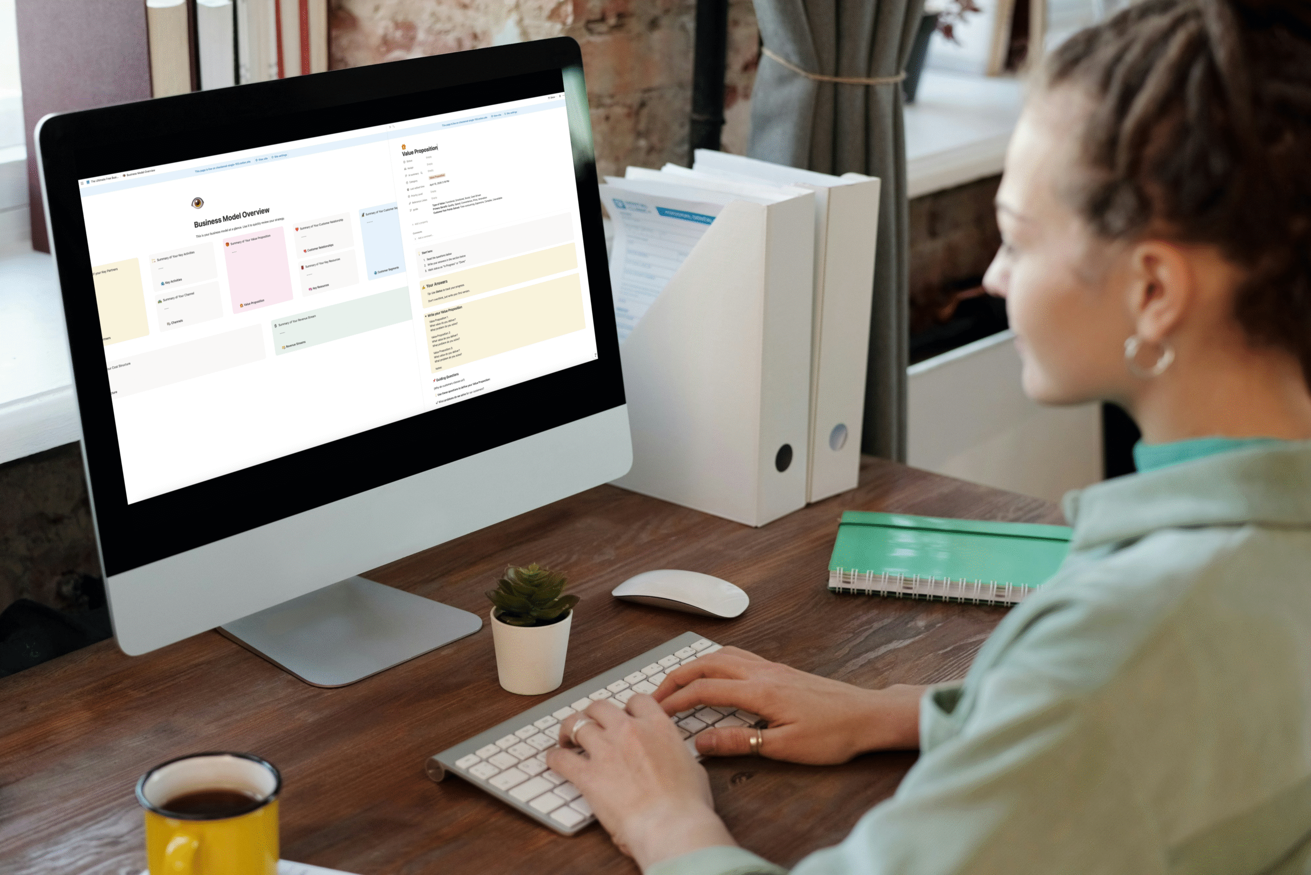 A person working at a desktop computer in a cozy workspace, viewing a Notion-style Business Model Canvas interface on the screen, with a keyboard, notebook, and coffee mug on a wooden desk near a window.