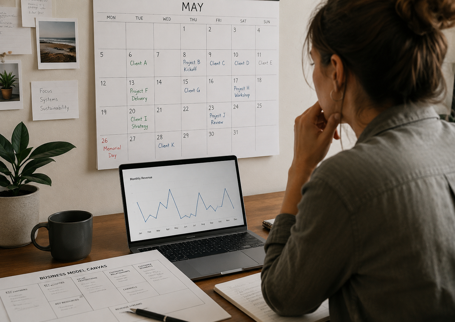 A small business owner sits at a desk looking at a laptop with an uneven monthly revenue chart, while a wall calendar filled with client projects and a Business Model Canvas worksheet suggest planning for more predictable income.